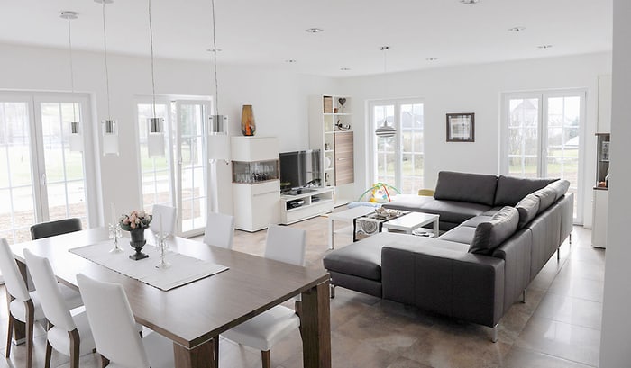 Modernes, offenes Wohnzimmer mit grauer Eckcouch, Esstisch und weißen Stühlen, großen Fenstern und Holzboden.